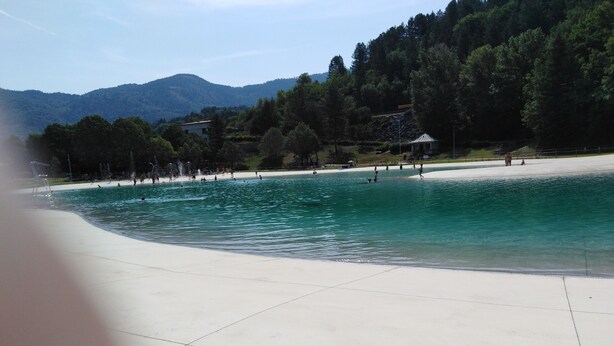 Ein Strand mit klarem Wasser, einem Pavillon und einer bewaldeten Hügelkette im Hintergrund.