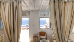 Una habitación con vista al mar, una silla de mimbre, una mesita con una vela y cortinas.