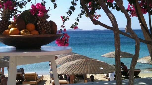Un espacio para comer frente a la playa, con una mesa preparada para dos personas, un frutero y vistas al mar.