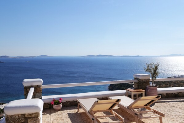 A terrace with two sun loungers, a small table, and a potted plant overlooking a clear blue sea with distant landforms.