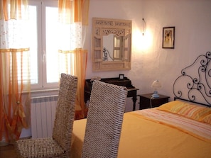 Une chambre à coucher avec un lit, une chaise, une commode, un miroir et une fenêtre avec des rideaux.