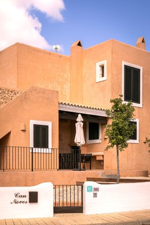 A building with a flat roof, a balcony with a table and chairs, and a small tree in front.