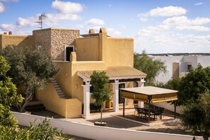Un edificio de estilo mediterráneo con fachada de piedra, un porche cubierto con mobiliario exterior y vistas al mar.