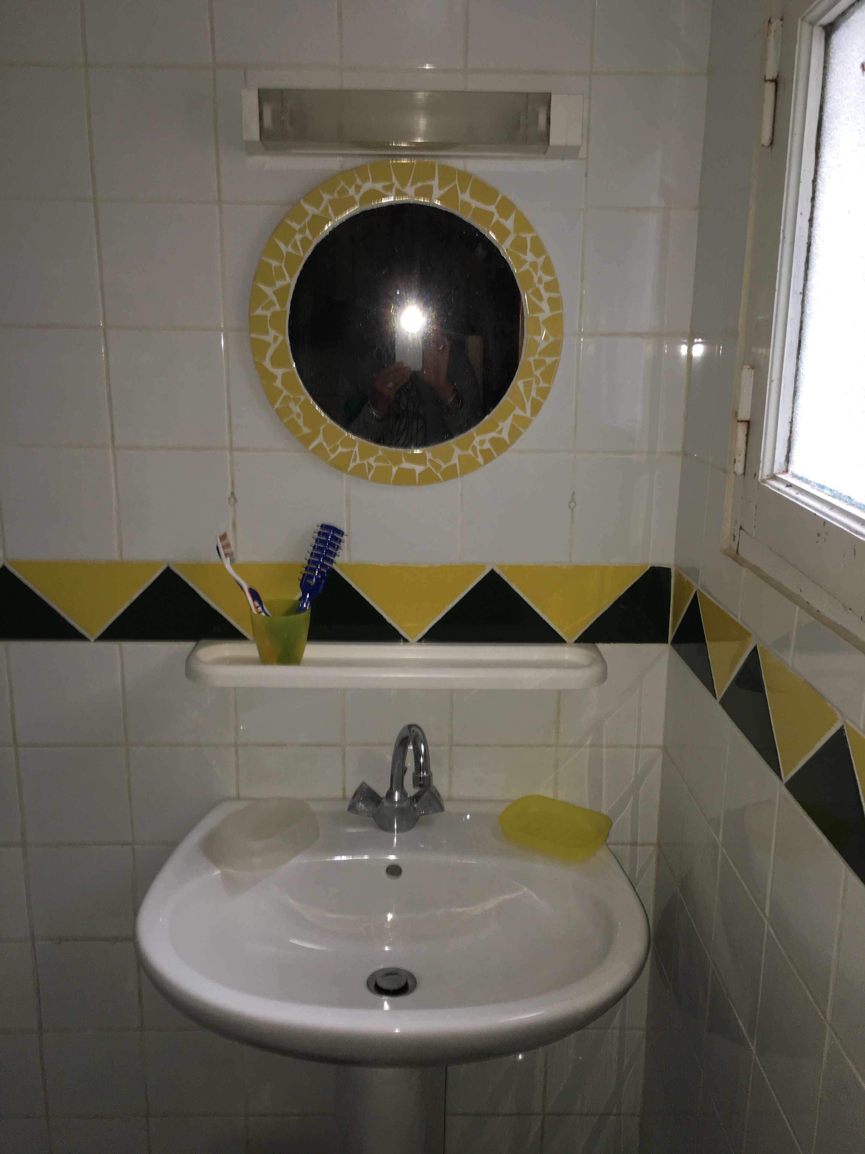 A bathroom with a white sink, a mirror with a yellow and white mosaic border, and a toothbrush holder with a toothbrush.