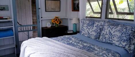 A bedroom with a bed, a nightstand, a shelf with books, a window, and a door.