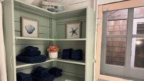 Um quarto com porta verde, janela com mosquiteiro e prateleiras com toalhas dobradas e objetos decorativos.