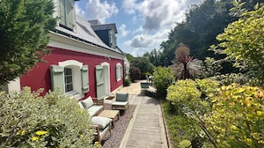A red building with a wooden deck, outdoor seating, and lush greenery. - House / Villa - Le Palais (Bangor)
