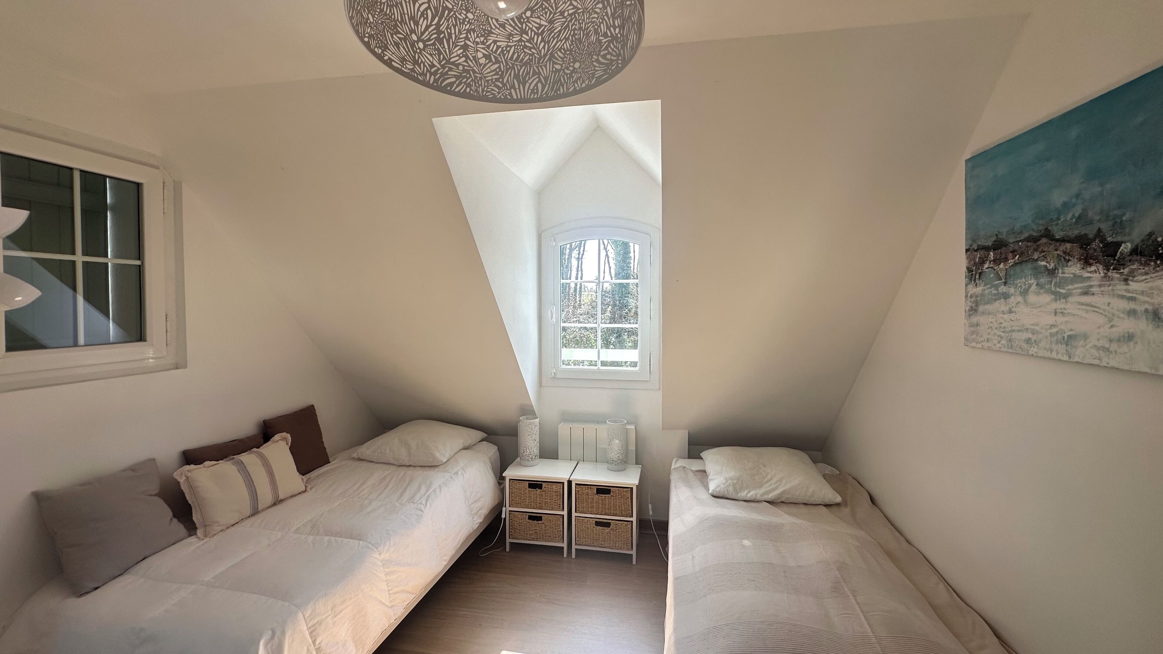 Ein Schlafzimmer mit zwei Einzelbetten, einem Fenster mit Blick auf Bäume, einer Deckenleuchte und einem Bild an der Wand.