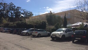 Ein Parkplatz mit mehreren Autos, Bäumen und einem Gebäude mit Satellitenschüssel im Hintergrund.