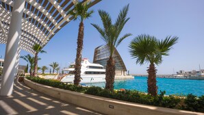 Una passeggiata lungomare con uno yacht bianco ormeggiato, palme e un edificio moderno con una struttura del tetto particolare.