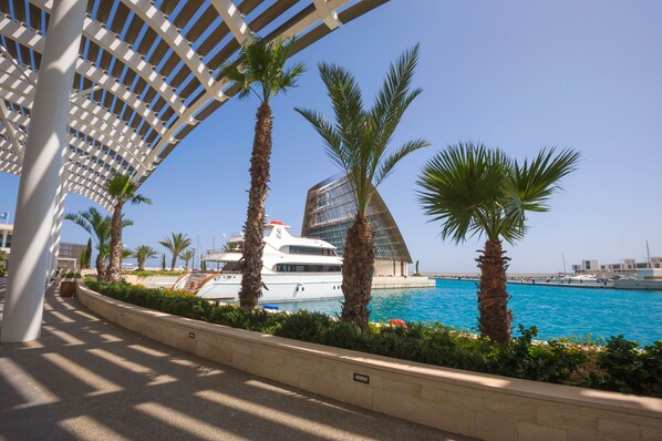 Une promenade en bord de mer, avec un yacht blanc amarré, des palmiers et un bâtiment moderne doté d’une structure de toit originale.