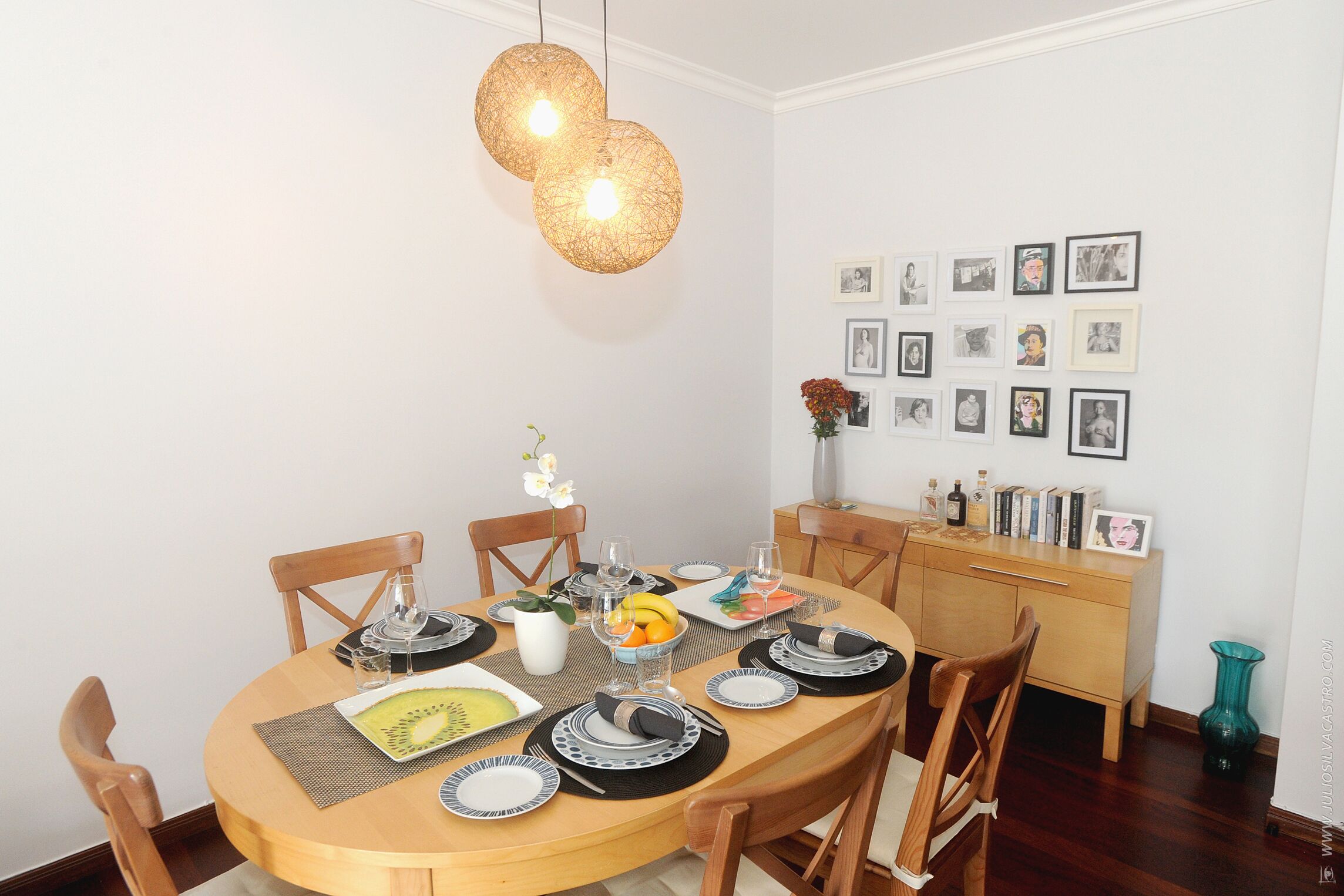 Een eethoek met een ronde tafel gedekt voor vier personen, omringd door houten stoelen. Daarboven hangen twee lampen en aan de muur hangen prestaties in lijsten.
