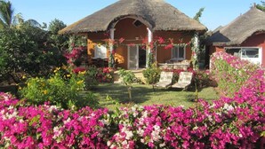 A bungalow with a thatched roof, surrounded by a garden with vibrant flowers and a clear blue sky.