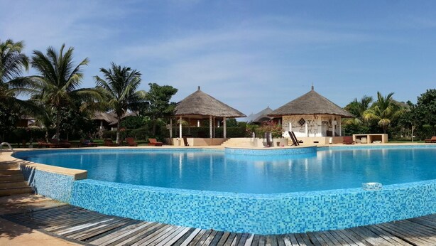 A large, clear blue pool with a wooden deck surrounding it, two thatched-roof structures, and palm trees in the background.
