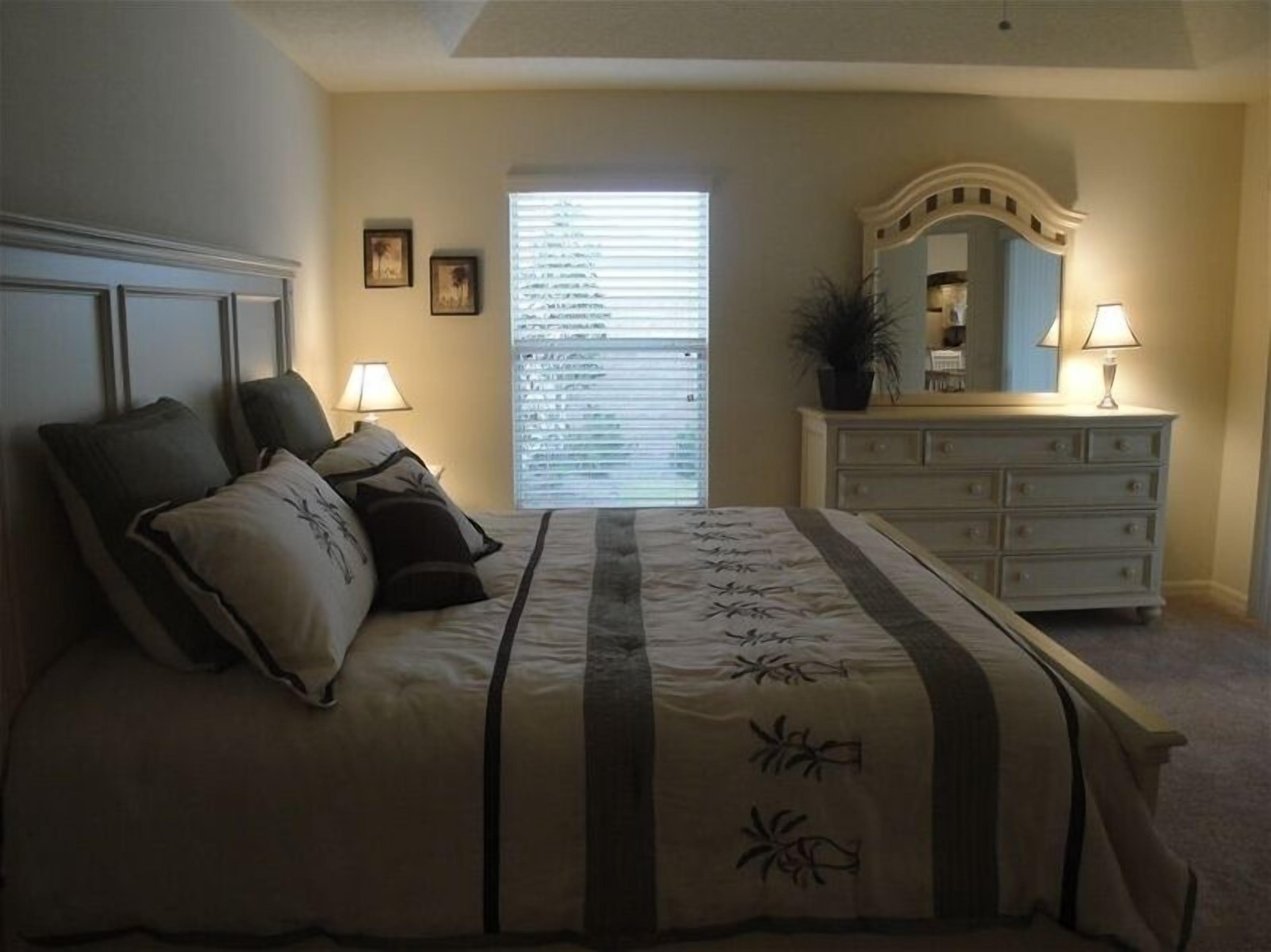A bedroom with a bed, two bedside lamps, a dresser, a mirror, and a window with blinds.