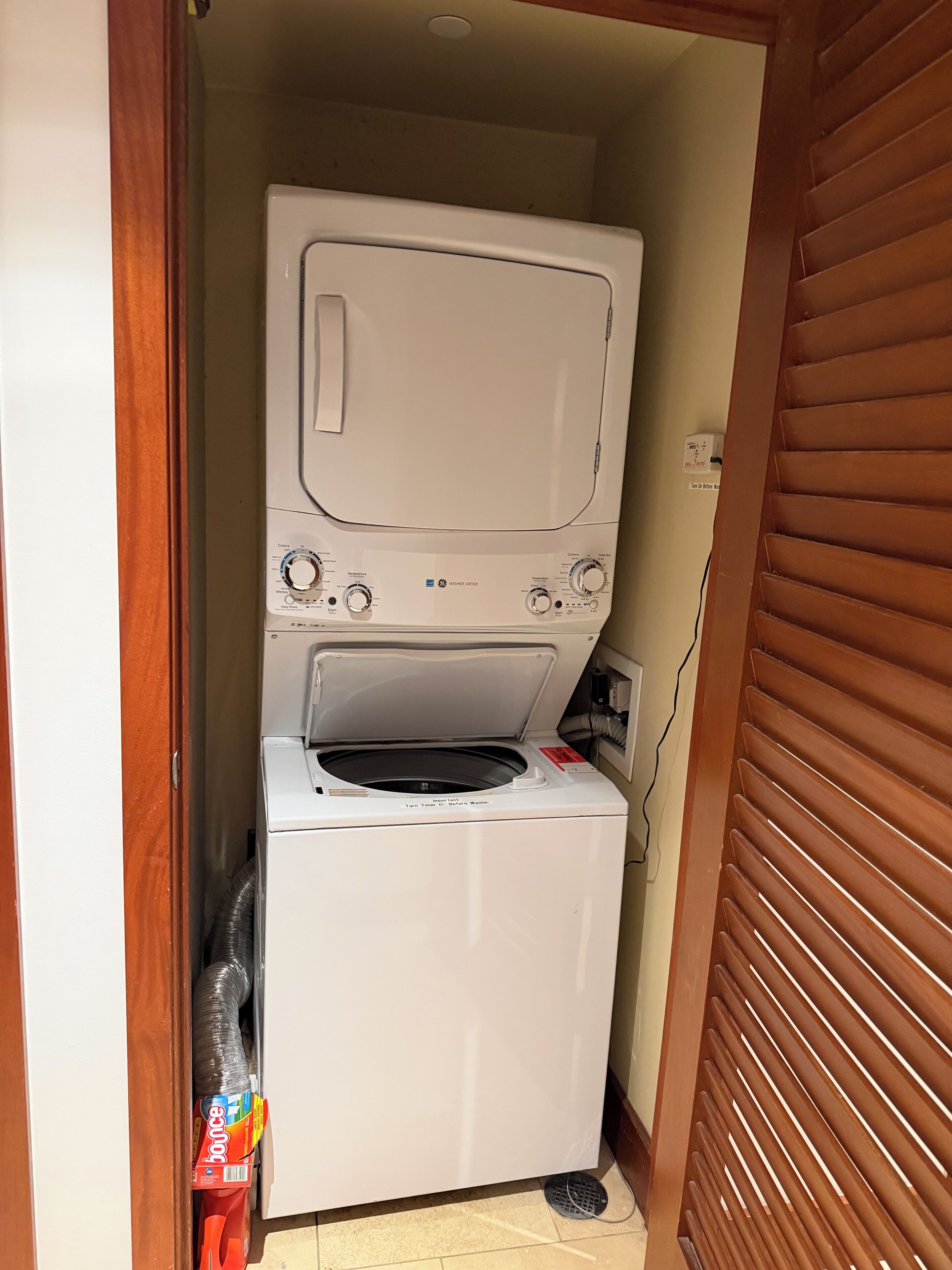 A compact washer and dryer unit in a small laundry room with a wooden door and a power outlet visible.