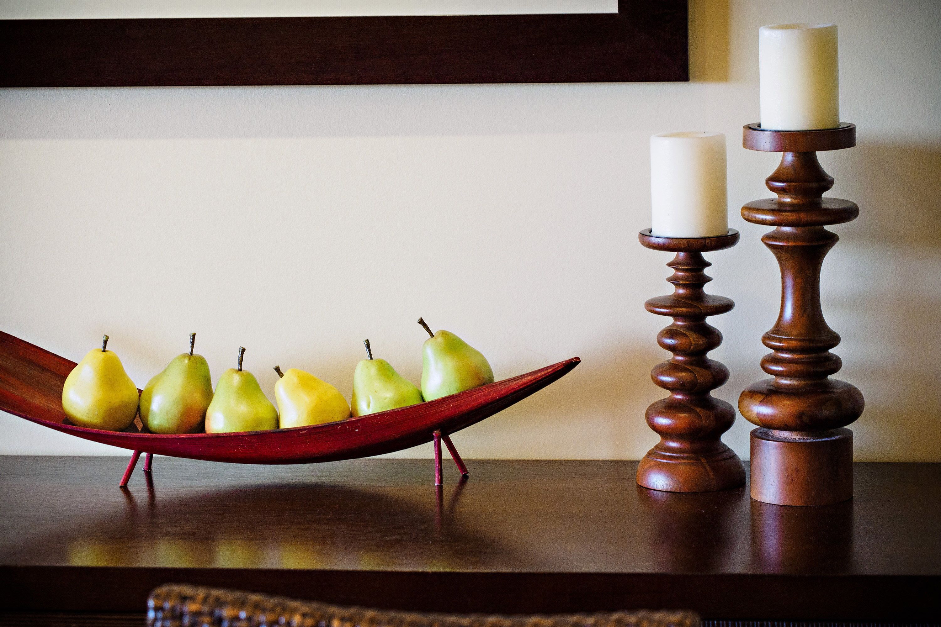 A wooden table with a red bowl containing pears, two candles on tall wooden stands, and a dark wooden surface.