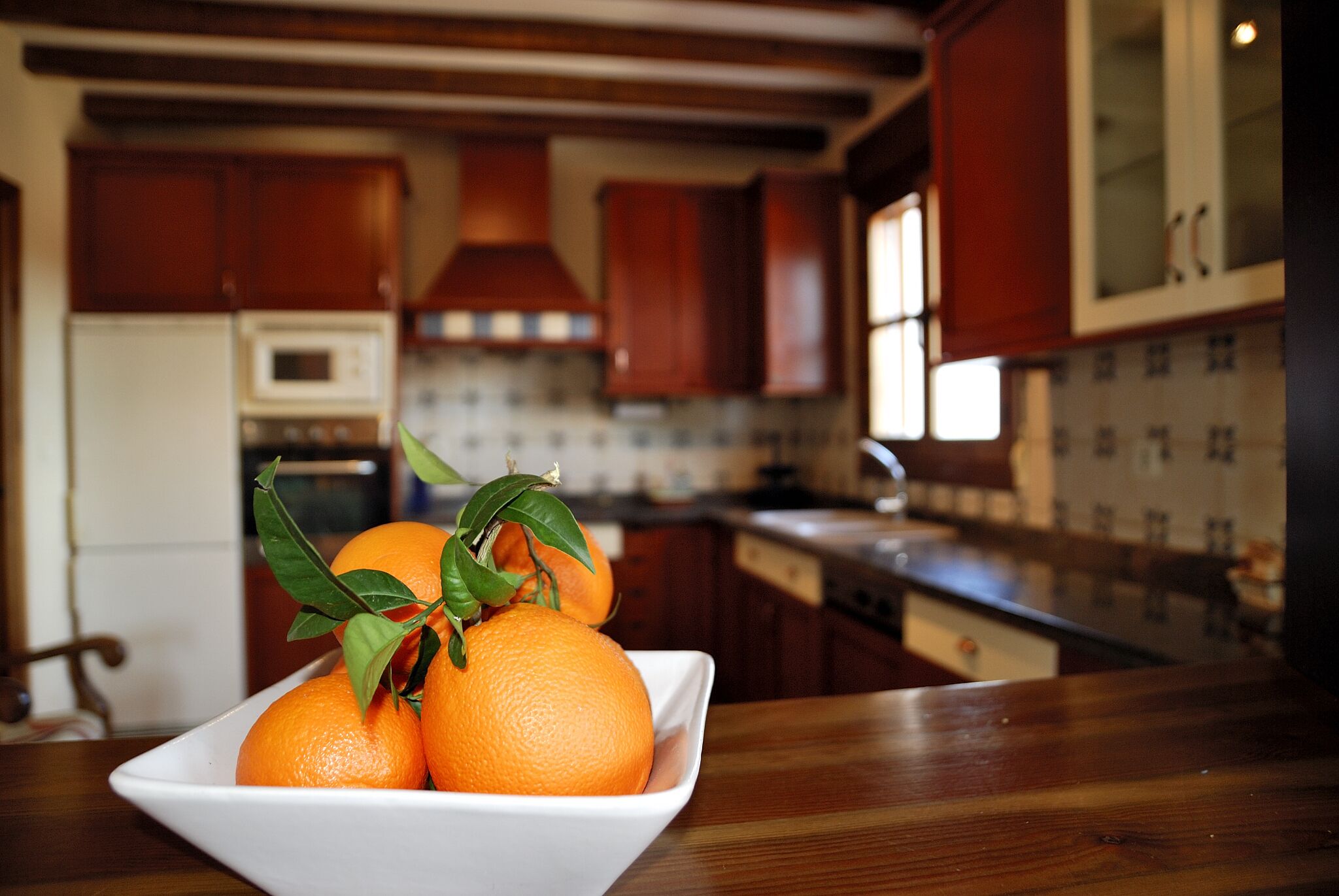Une cuisine avec des meubles de rangement en bois, un plan de travail blanc et un saladier rempli d’oranges.
