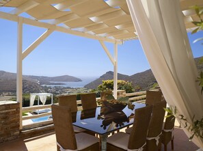 A covered outdoor seating area with wicker chairs and a glass table, overlooking a scenic landscape with hills and a body of water.