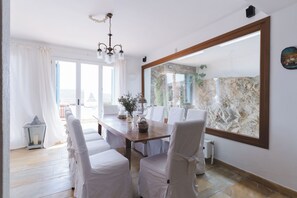 Een eetruimte met een grote spiegel, witte stoelen, een houten tafel en uitzicht op het buitenleven door een glazen deur.