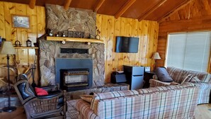 Interior acogedor de una cabaña con chimenea de piedra, paredes de madera, un sofá de cuadros y un sillón de cuero.