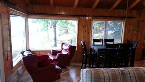 Interior de una cabaña de madera con una mesa de comedor, dos sillones de cuero rojo, un sofá de cuadros y ventanas amplias que ofrecen vistas al exterior.