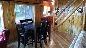 Interior de una cabaña de madera con una mesa de comedor, sillas, un sofá y una escalera.