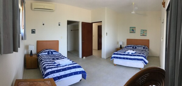 A hotel room with two beds, a wooden nightstand, a small table, and a chair. There is an air conditioning unit on the wall, a door leading to another room, and a ceiling fan. - BEROCO Front Beach House Yucatan Wi-Fi, Pool, Smart Tv (Uaymitun)