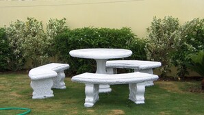 Un espace extérieur aménagé avec des sièges blancs, comprenant une table ronde et des bancs, le tout entouré de verdure et adossé à un mur jaune.