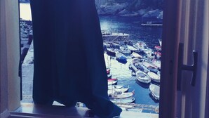 Une vue sur un port avec des bateaux amarrés, des bâtiments en arrière-plan et une personne debout près d’une fenêtre.