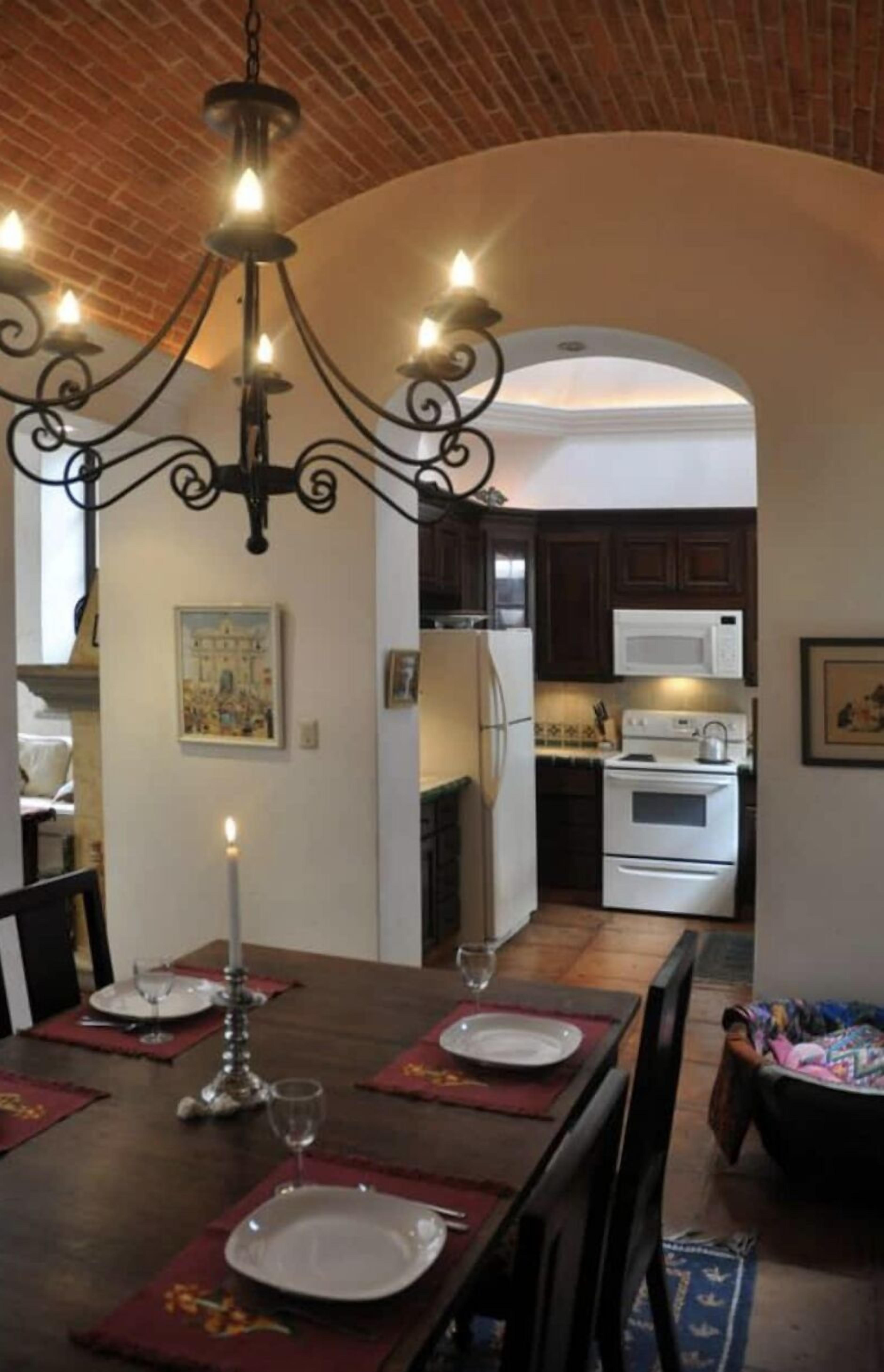 Comedor con candelabro, una mesa dispuesta para cuatro personas y vista a la cocina.