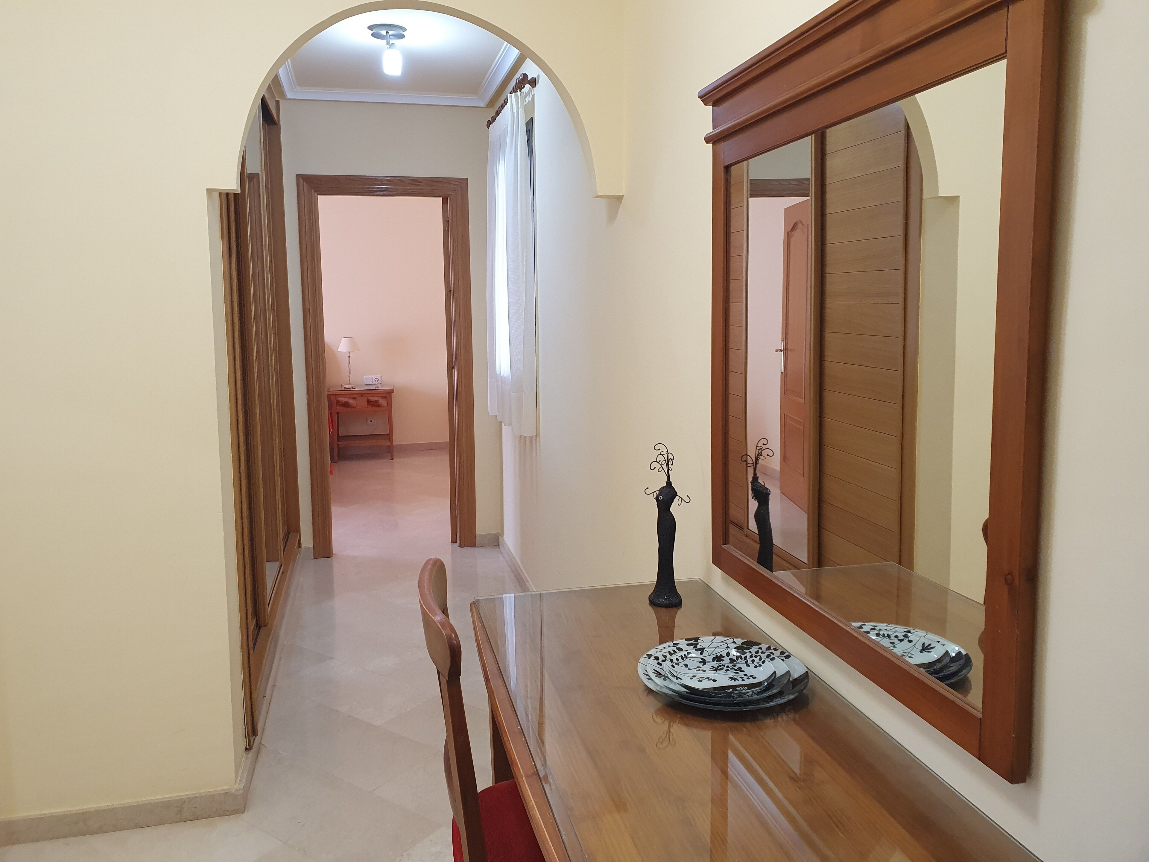 Un pasillo con una mesa de comedor de madera, sillas, un espejo y una mesita con una lámpara y un radio.