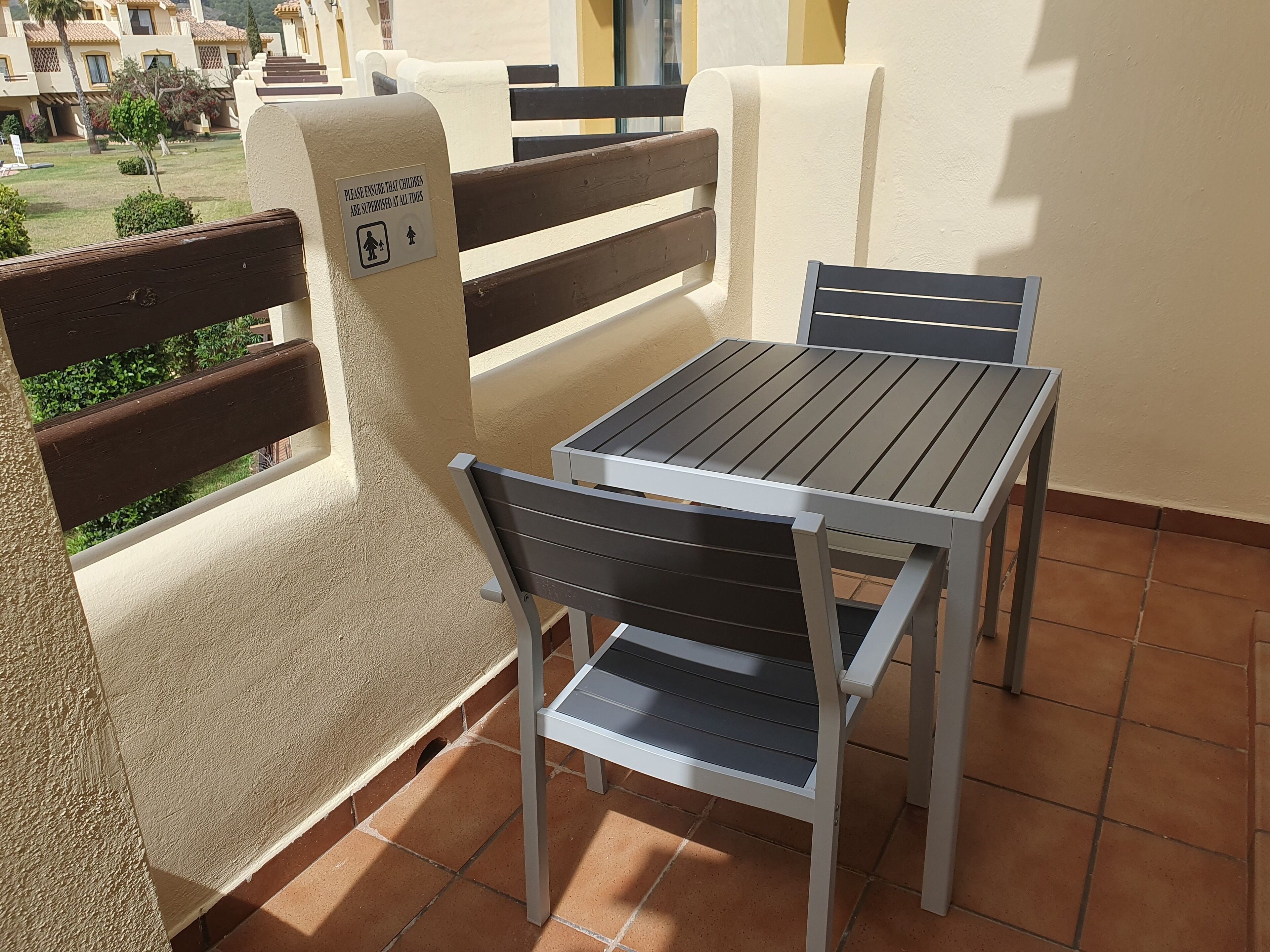Un balcón con mesa y sillas, un cartel que indica supervisión infantil y vistas a un jardín y edificios.