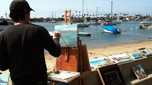 Un hombre pinta un paisaje sobre caballete junto a la orilla, con barcos y palmeras al fondo.