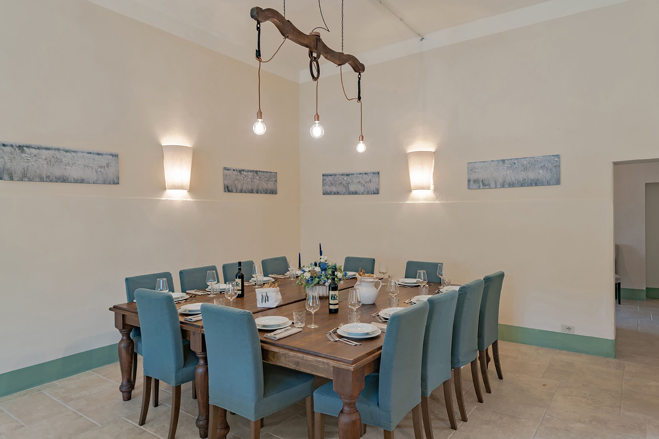 Ein Essbereich mit einem Holztisch, gedeckt für sechs Personen, umgeben von blaugrünen Stühlen, mit an der Wand befestigten Lampen und abstrakten Gemälden.
