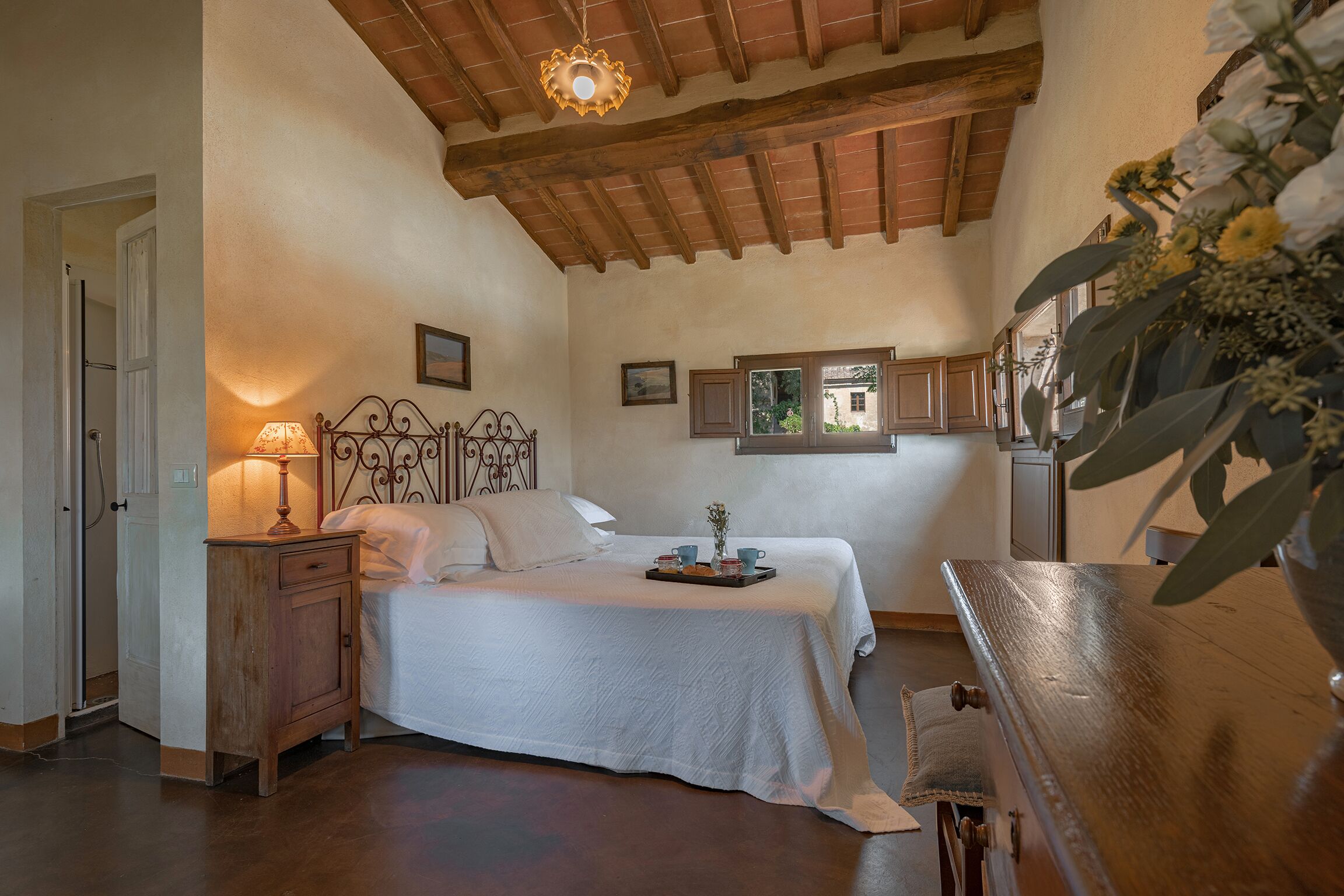 Une chambre avec un plafond en bois, un lit recouvert d’une courtepointe blanche, une table de chevet avec une lampe et une fenêtre donnant sur un paysage verdoyant.