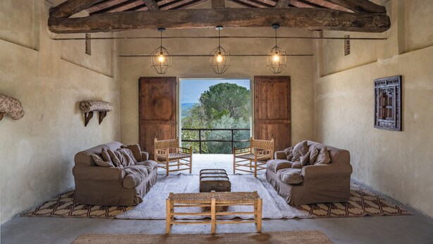 A rustic room with a high ceiling, wooden beams, and a large window offering a view of trees. There are two sofas and two wooden chairs arranged around a central bench. - Luxury Ecofriendly Country Villa  - Val d'orcia (Buonconvento Siena)
