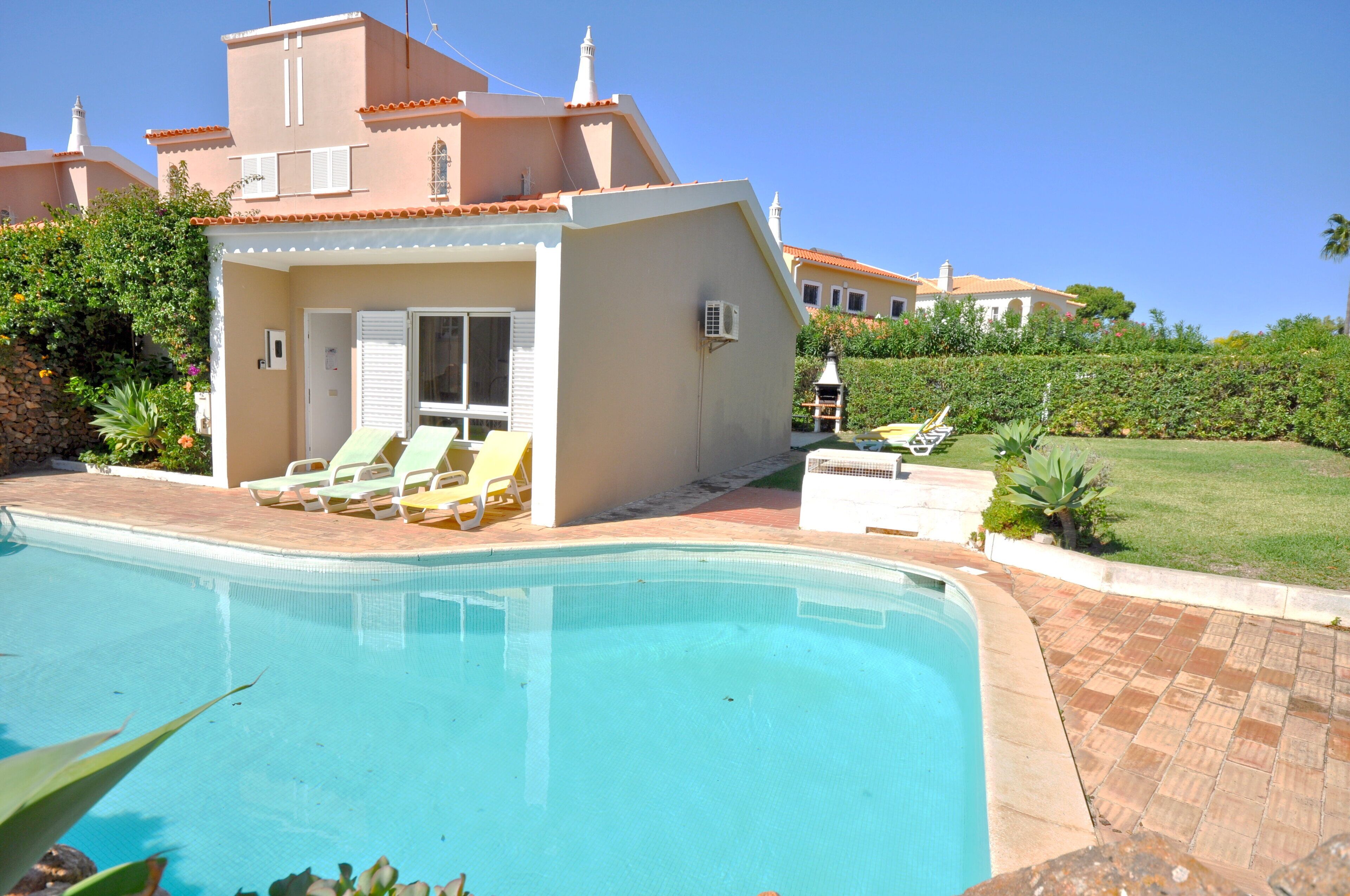 A pool area with lounge chairs, a small building with a tiled roof, and a well-maintained lawn.