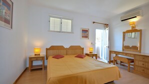 Quarto com uma cama grande, duas mesas de cabeceira com luminárias, um aparador de madeira, um espelho e uma janela com persianas.