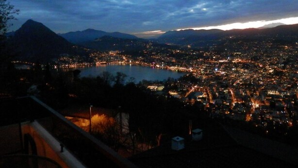 Nachtansicht einer Stadtlandschaft mit See, Bergen und Blick von einem Balkon.