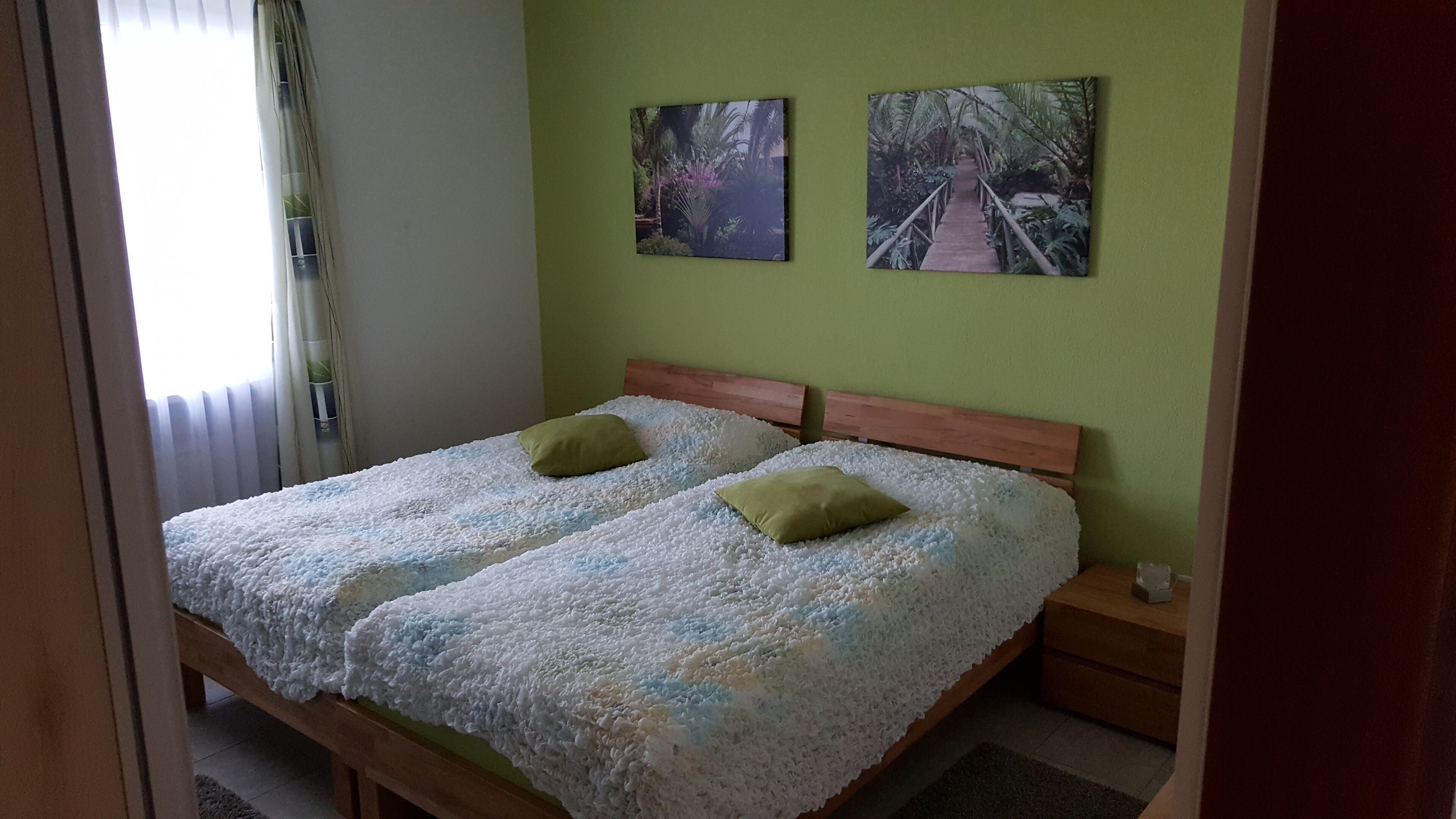 A double bed with a textured white blanket and two green pillows, a wooden headboard, and a small bedside table with a lamp.