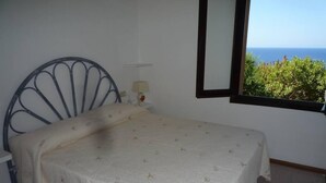 A single bed with a metal headboard, a bedside lamp, and a window with a view of greenery and the sea. - Costa Paradiso: Two-room apartment with stunning views (Costa Paradiso)