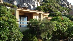 A small, single-story building with a balcony, surrounded by lush greenery and rocky terrain. - Costa Paradiso: Two-room apartment with stunning views (Costa Paradiso)