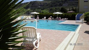A swimming pool with lounge chairs and umbrellas, surrounded by greenery and a hillside in the background. - APARTMENT WITH POOL (Pizzo)