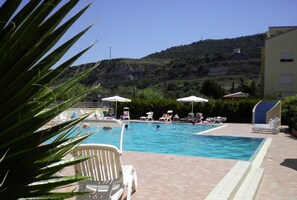 A swimming pool with lounge chairs and umbrellas, surrounded by greenery and a hillside in the background.