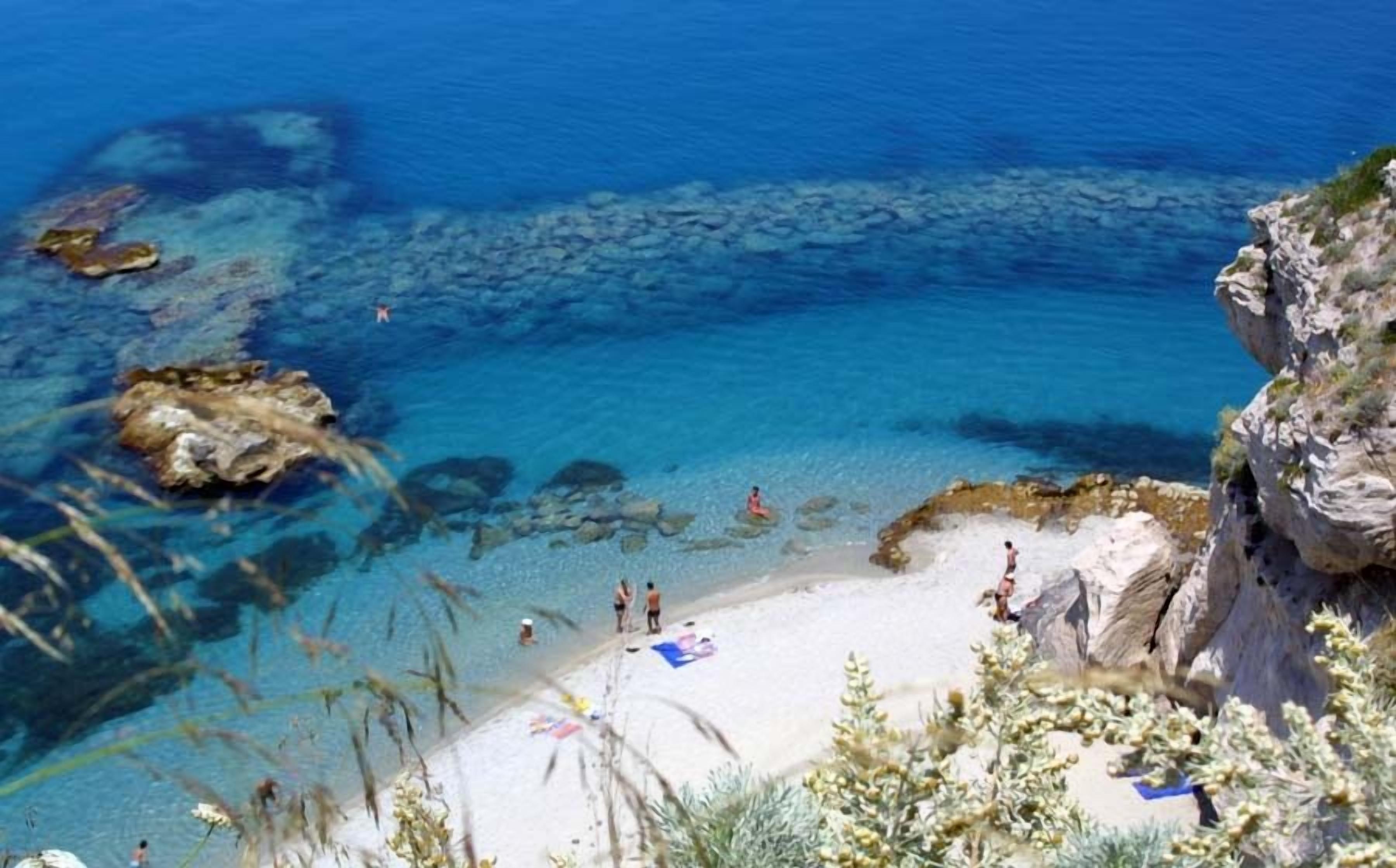 Une plage côtière aux eaux bleues et limpides, parsemée de formations rocheuses, où les gens profitent du soleil et de la mer.