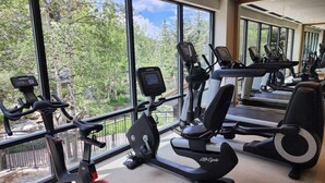 Un gimnasio con diverso equipo de ejercicio, amplias ventanas y vista a árboles.