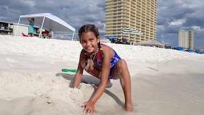 Una spiaggia con un bambino che gioca sulla sabbia, una tenda bianca e un edificio alto sullo sfondo.