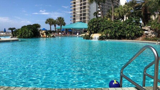 Una piscina cristallina blu con una scaletta in metallo, circondata da palme e un grande edificio con balconi.