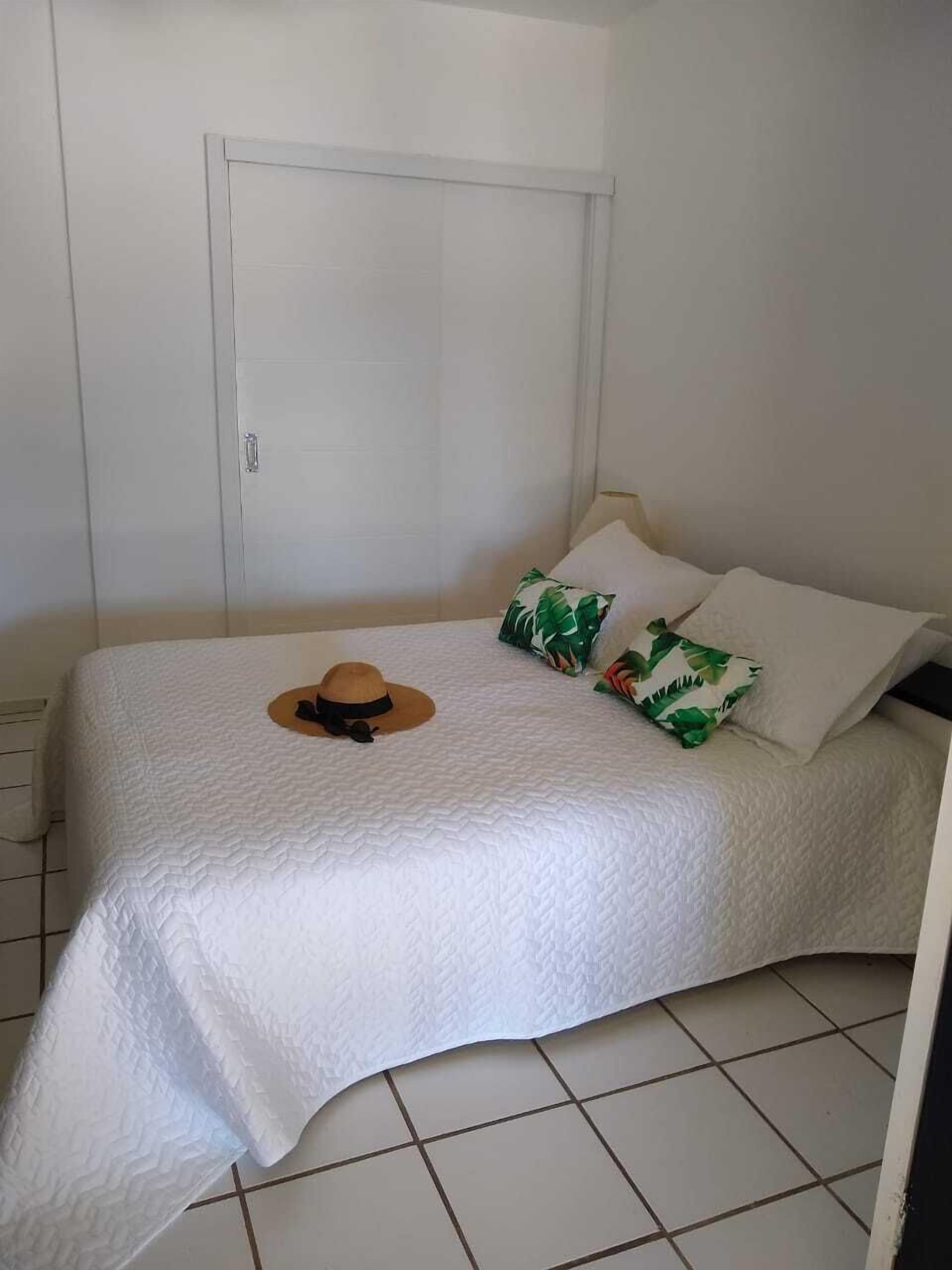 Une chambre avec un lit simple, recouverte d’une courtepointe blanche matelassée, une porte en bois et un sol carrelé.